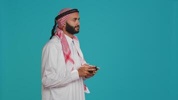 Modern man playing games with controller, having fun with video gaming competition over blue studio background. Islamic player in traditional arab clothing competes in tournament. photo