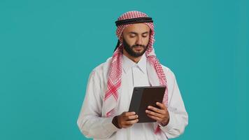 Middle eastern guy browsing tablet on camera, navigating webpages online on digital device. Male model in arabic clothing using gadget to check internet sites, texting on social media. photo