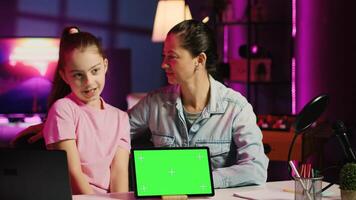 Cheerful child and loving mom record chroma key tablet review on online platforms. Young smiling daughter and parent presenting isolated screen device, filming in home studio with pink neon lighting photo