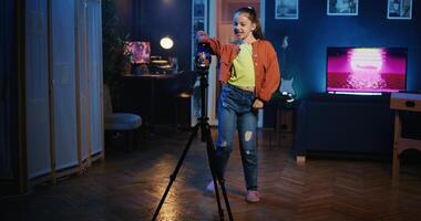 Dolly in shot of kid media star recording trendy dance video clip for social media using cellphone on tripod stabilizer, entertaining audiences. Gen z influencer recording content using smartphone photo
