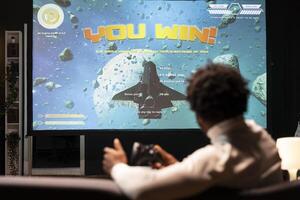 Man looking at winning screen of science fiction game on large smart TV. African american gamer receiving victory message after completing all videogame levels, watching statistics roll on display photo