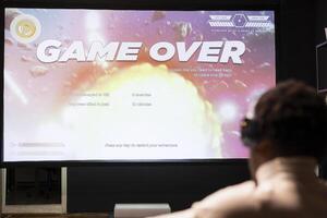 Gamer wearing headphones upset at seeing game over message on widescreen TV while playing arcade space shooter videogame. Man relaxing at home on gaming system, disappointed after losing photo