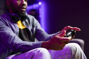 Gamer on couch using controller and headphones to play videogame at home, close up. African american man in home theatre using high tech gaming console gamepad to defeat opponents in game photo