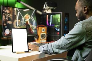 Mockup tablet on desk next to BIPOC man playing SF videogame on computer, talking into headset with friends. Isolated screen portable device and gamer using headphones to chat with team in game photo
