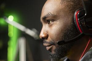 Smiling BIPOC man has fun playing videogames on PC while chatting with teammates through headphones mic. Close up of upbeat african american gamer communicating with friends through headset microphone photo