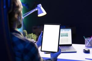 Man vertically holding tablet with isolated white screen in front of his laptop. Male freelancer working from home, focused on digital device displaying blank chromakey mockup template. photo