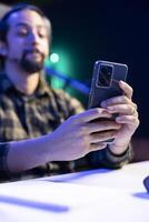 Selective focus of cell phone being held by male adult seated at the table, surfing the net and answering text messages. Young guy looking at social media platforms on his mobile device. photo