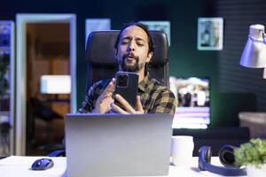 Dedicated male individual engrossed in an interesting video call on his smartphone, conversing with family and friends. Young guy grasping cell phone and speaking in a virtual meeting. photo