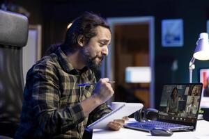 During a video call, young male college student smiles while comparing notes with his classmates on his laptop. He holds a notebook in his hand and conducts a discussion in a virtual meeting. photo