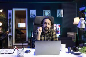 Portrait of young man sitting in front of his laptop at his home desk, working from home. Serious male individual holding a pen while looking at the camera, preparing for work and research. photo