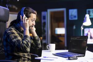 Male freelancer working at a desk focused on his laptop that is displaying code and data. Man wearing wireless headphones is checking software information and ensuring cyber security. photo