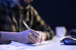 Person studying and researching information while writing on a notepad at a table. Closeup of male individual making notes with a pen on paper, organizing their schedule, and finishing tasks. photo