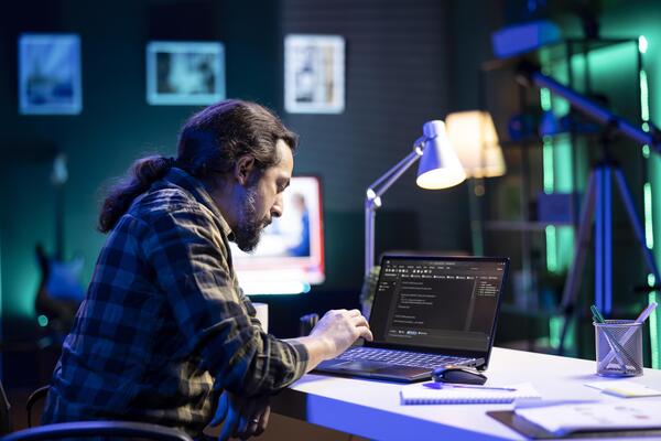 Dedicated male programmer uses his portable computer to manage databases and keep an eye on network security. Bearded man is coding on a digital laptop while seated at a desk.
