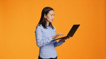 Casual person navigating online on laptop in studio, looking for inspiration on wireless pc against orange background. Asian woman browsing webpages on internet, search project. photo