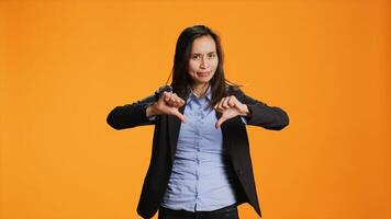 Asian employee showing dislike sign in front of camera, presenting thumbs down with unhappy facial expressions. Young adult does rejection and disagreement symbol over orange backdrop. photo