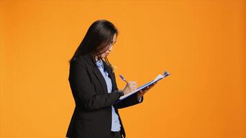 Asian manager writes important data on papers with clipboard, taking notes to create full report in front of orange background. Young woman in business clothing filling in documents, keeping track. photo