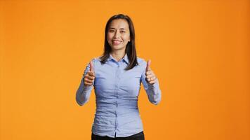 Asian model presenting thumbs up symbol in studio, expressing good vibes and success against orange background. Young smiling person giving like sign on camera, shows approval gesture. photo