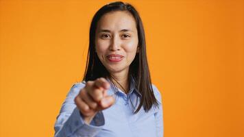 contento asiático persona señalando dedo a cámara durante disparar tiempo. positivo joven mujer con casual actitud utilizando carisma, en pie en estudio terminado naranja fondo, demostración algo. foto