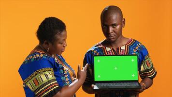 Ethnic couple pointing at laptop with greenscreen display, posing in studio with portable pc presenting isolated copyspace layout. Man and woman looking at blank mockup template on camera. photo