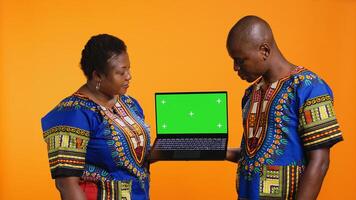 Ethnic people presenting laptop with greenscreen layout, showing blank mockup chromakey display and posing in studio. African american husband and wife pointing at isolated copyspace. photo