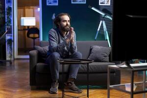 Hopeful man praying for favorite team to win while watching live sports event broadcasted on TV. Supporter sitting on couch at home doing imploring hand gesture, enjoying football competition photo