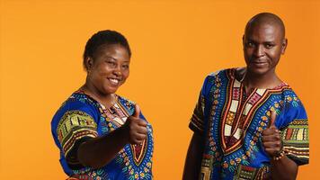 Joyful husband and wife doing thumbs up sign in studio, presenting their approval and satisfaction. African american couple showing like symbol and confirming good feedback on camera. photo