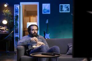 Person using TV to binge series on subscription based streaming services, enjoying bowl of popcorn. Man relaxing in apartment watching VOD shows on television display, eating snack photo