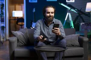 Smiling man watching shows on TV, using remote control, texting friends on smartphone. Cheerful person in pajamas consuming mindless entertainment and chatting with mates on phone photo