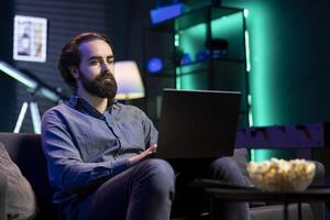 Man sitting in cozy apartment on couch typing on laptop keyboard, watching TV with bowl of popcorn ready. Teleworking person solving tasks on notebook with film playing in background photo