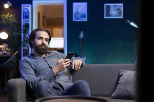 Man lounged on couch texting friend on smartphone, inviting him to watch movie on streaming service. Person at home discussing with mate on mobile phone, recommending TV show photo