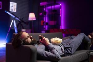 Gamer lounged on couched while engaging in online competitive multiplayer tourney using gaming console and gamepad. Man in home theatre playing videogames with bowl of popcorn in lap photo