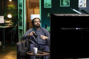 Happy man playing shooter videogame in neon illuminated home, holding controller. Gamer excited to participate in online tournament, battling other players using professional joypad photo