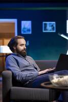 Smiling man enjoying bowl of popcorn while watching TV and solving tasks on laptop. Spectator on couch in living room eats snacks during movie and scrolls on social media using notebook photo