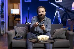 Portrait of smiling man playing videogame and eating snacks, holding controller. Cheerful gamer participating in esports tournament using professional gamepad for better victory chances photo