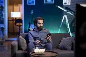 Person in home theater binging series on streaming service, texting friend on phone. Man in neon lit apartment with bowl of popcorn in front watching shows on television and chatting with mate photo