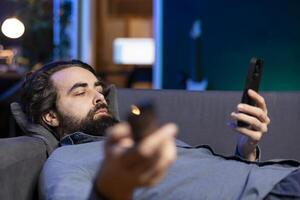 aburrido persona descansado en sofá atracones serie en transmisión servicios, hojeada utilizando remoto y mensajes de texto en teléfono. hombre acecho vod muestra en televisión, zapping aunque canales, chateando en teléfono inteligente foto