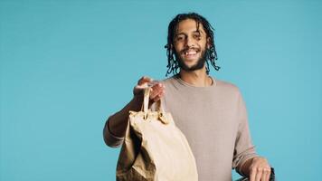 Portrait of pizza delivery guy taking takeaway paper lunchbag out of thermal bag. Middle Eastern courier getting meal out of food backpack, isolated over blue studio background, camera A photo