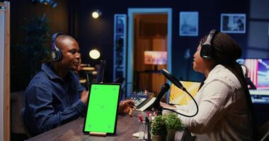 Isolated screen tablet next to vlogger show host chatting with guest in neon lights ornate living room studio. Chroma key device on table with man and woman sitting and discussing on podcast photo