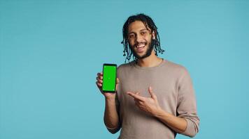 Excited man pointing index finger towards isolated screen phone, doing recommendation. Joyous person presenting mockup smartphone, doing positive gesturing, blue studio background photo