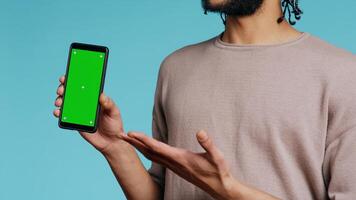 Happy man pointing towards green screen mobile phone, showing thumbs up sign. Cheerful person presenting chroma key cellphone, doing gesturing, isolated over studio background, camera B close up photo