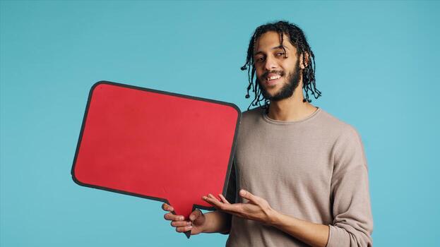 Smiling BIPOC man holding red speech bubble sign of empty copy space for message. Joyous person presenting thought bubble cardboard used as promotion concept, isolated over studio background photo