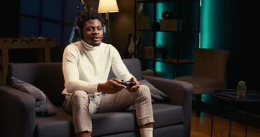 BIPOC man playing shooter videogame in neon lit apartment, holding controller. Gamer participating in internet multiplayer game using console system and headphones, zoom out shot photo