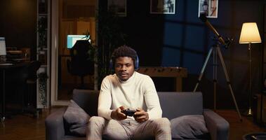 African american man playing shooter videogame in warm apartment, holding controller. Gamer participating in PvP online multiplayer game using console system and headphones for immersive experience photo