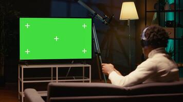 African american man sitting on couch at home, relaxing by playing videogames on green screen TV. Gamer unwinding in apartment by competing in online multiplayer game displayed on mockup television photo