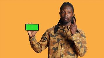 Cheerful adult holds smartphone with greenscreen in studio, presenting isolated copyspace display on mobile phone layout. Young person showing blank chromakey mockup on device. photo