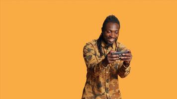 Joyful person playing online videogames on mobile phone, having fun with gaming tournament on device in studio. African american man enjoying shooting game with friends, orange background. photo