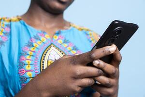 African american woman using mobile phone to text and browse the internet, navigating social media and web technology. Young model wears ethnic colorful costume over blue background. photo