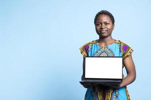 African american person does advertisement with white screen on laptop, presenting wireless computer that runs blank isolated layout. Young woman smiling at camera and holding pc. photo