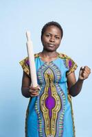 African american displeased person using rolling pin to threaten someone in front of camera, wearing traditional historic outfit. Young serious adult being aggressive, starting a fight. photo