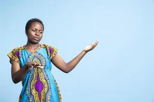 African american woman doing an advertisement by pointing at left or right sides, working on new promotional ad in studio. Young model in traditional colorful outfit smiling on camera. photo